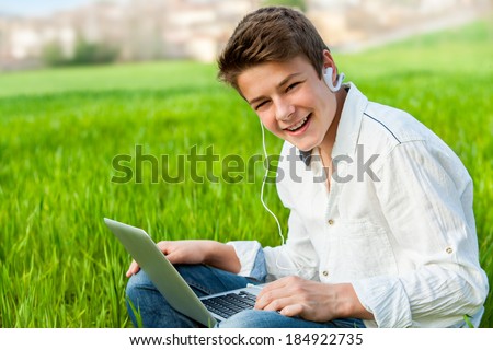 Portrait of handsome teen student working on laptop in green grass field.