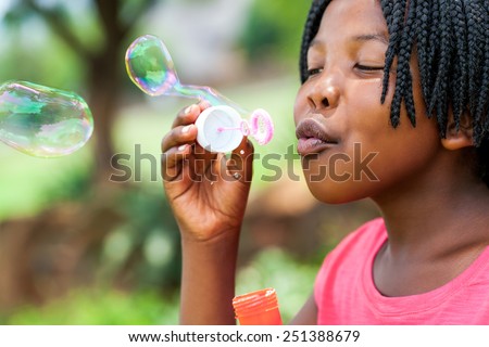 Close up portrait of cute African girl with braids blowing bubbles in park.