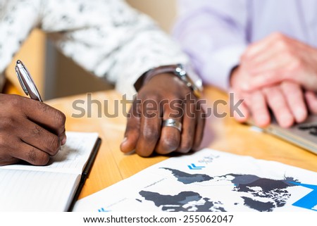 Close up detail of black business man signing documents with caucasian colleague in background.
