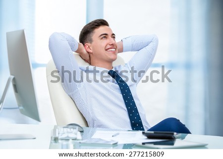 Close up portrait of young office worker daydreaming at desk. Young man sitting at desk with hands behind head and looking up.