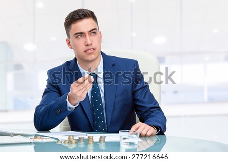 Close up portrait of young Businessman at desk holding money coins with wondering face expression. Young businessman in blue suit sitting at desk in office looking up.