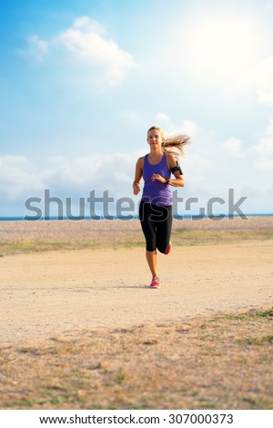 Attractive young woman running with smart watch and armband along beach in early morning sun.