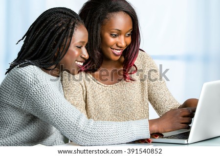 Close up portrait of two attractive african teen girls sitting together at table. Girl with braided hair pointing at computer screen.