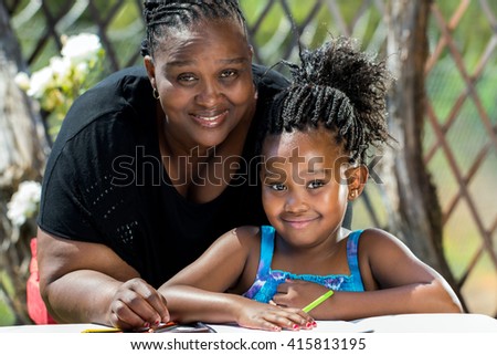 Close up portrait of african mother and little daughter with braided hairstyle in garden.Girl drawing on paper with color pencils outdoors.