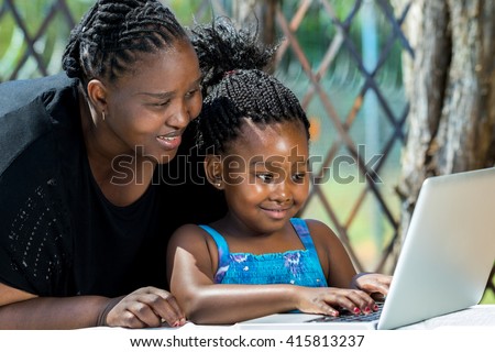 Close up portrait of African mother and little girl with braided hairstyle looking at laptop. Cute girl typing on keyboard at table in garden.