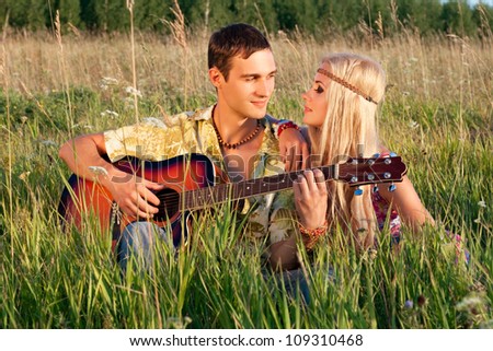 cute young man and fine woman sit on meadow. young man plays  guitar. style of hippie