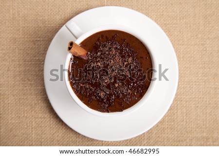 hot chocolate in white mug with cinnamon stick, saucer and 100 percent chocolate flakes, on jute backdrop, top view