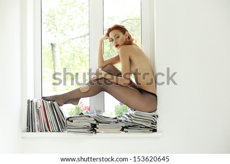 Auburn Nude Woman in Torn Tights on Magazines over Window Sitting. Glamor