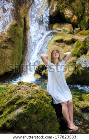 Beautiful young woman in white sundress and straw hat stopped to rest and dream near the waterfall among the mossy stones
