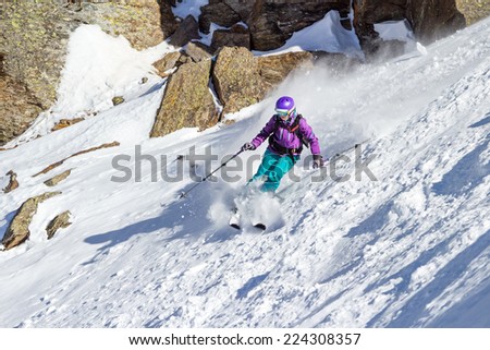 Woman skier at a steep slope in deep snow in a turn raises the snow dust on the background of rocks