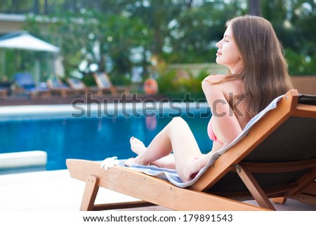 Happy young woman in bikini laying on chaise-longue luxury pool side