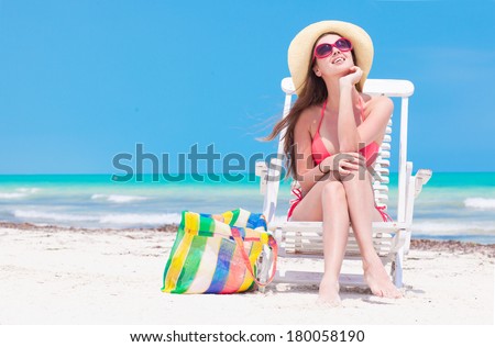 Woman in bikini and straw hat with beach bag sitting on chair on beach. back view