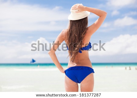 back view of beautiful young woman in blue bikini and straw hat standing on tropical beach enjoying the sun