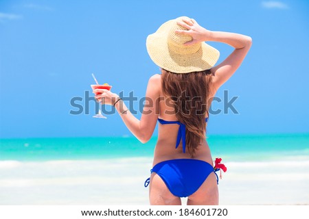 closeup back view young long haired woman in blue bikini with cocktail in hand and straw hat on beach