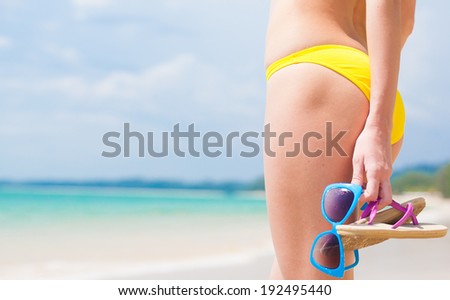 back view of woman in yellow bikini holding sunglasses and flip flops in her hand