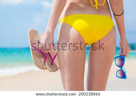 back view of woman in yellow bikini holding sunglasses and flip flops in her hand
