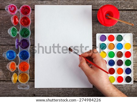 hand with brush painting with watercolor  and blank white paper sheet  on vintage wooden background