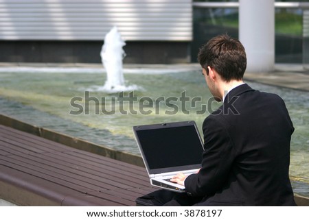 business man with laptop outside on a bench next to fountain working
