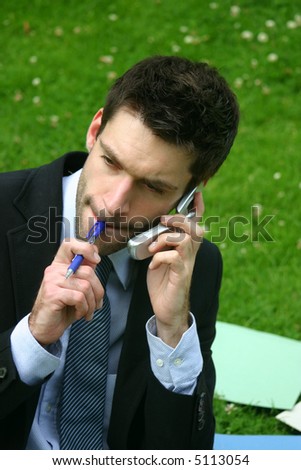 business man with designer stubble in the park having a conversation on cell phone while he\'s chewing on his blue pen