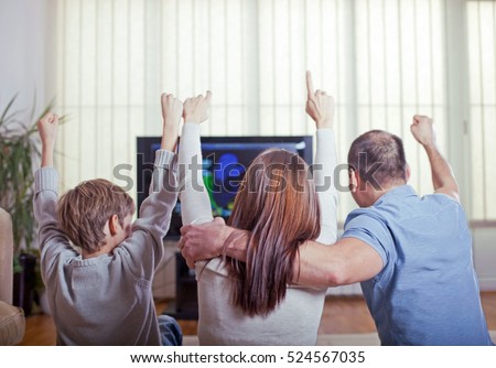 Family of three watching sports on TV and cheering with raised arms. Rear view.
