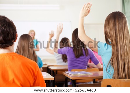 Rear view of students in bright classroom responding to the teacher\'s question, raising their arms up.