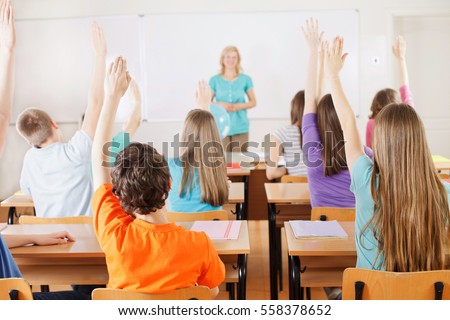 Rear view of students in bright classroom responding to the teacher\'s question, raising their arms up