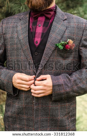The young man wears a gray jacket and buttoning.Cool Boutonniere on hip trendy groom at wedding. Grey suit, colorful vinous boutonniere. Maroon Bow tie.