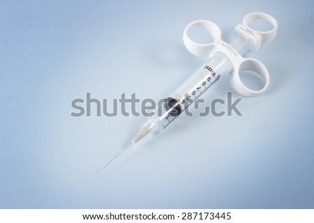 A Medical Syringe with needle sits on a light blue paper background.  The needle points toward the camera.