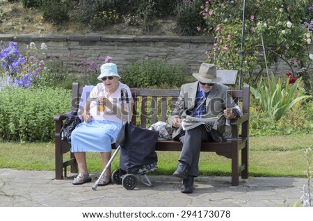 Clacton-on-sea, Essex, England. UK 16 June 2015-Happy elderly couple sitting on bench in the park, the man is reading the newspaper, the woman is drinking.