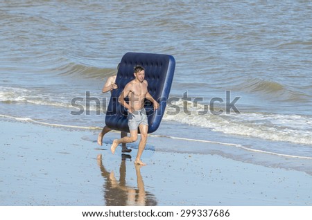 Clacton-on-sea, Essex, England, UK: 19 July 2015- two young men running along the beach carrying an inflatable airbed, to use in the sea.