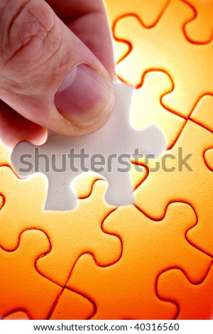 Fingers holding jigsaw puzzle piece over orange puzzle.
