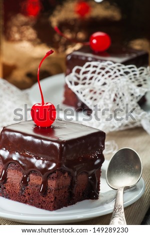 Two chocolate coated fudge cakes with candied cherries in white porcelain plate, with white lace and a golden background