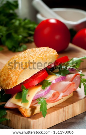 Gourmet sandwich with ham, cheese, fresh tomatoes, salad and a white bread bun, on a wooden cutting board and salad leaves in the background