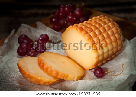 A round of cheese placed on packaging paper with red grapes on a wooden cutting board in a dark corner