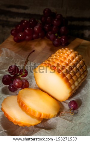 A round of cheese placed on packaging paper with red grapes on a wooden cutting board in a dark corner