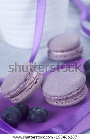Violet French macaroons cookies and blueberries on violet fabric with violet ribbon and white vase in background
