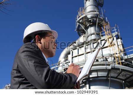 Industrial engineer standing in front of a large oil refinery machine, writing  under  pipes and cooling towers