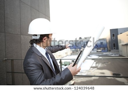 young architect with white helmet holding blueprint and clipboard pointing at the building construction