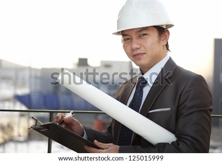 close-up young asian architect with white helmet holding blueprint and clipboard looking on camera