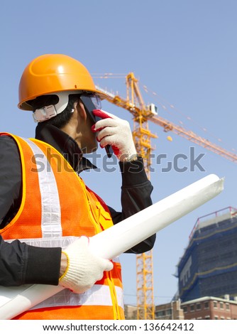 construction worker talking in mobile phone. Wearing orange safety vest and looking yellow crane on the background
