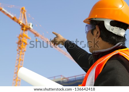 Engineer checking work plan on location site. Horizontal image asian engineer wearing orange safety vest and helmet under construction checking plan with yellow crane on the background and blue sky