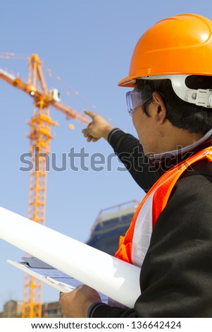Engineer checking work plan on location site. Vertical image asian engineer wearing orange safety vest and helmet under construction checking plan with yellow crane on the background and blue sky