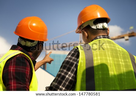 Two construction worker wearing safety vest discussion looking and pointing yellow crane at construction site Two construction worker wearing safety vest discussion looking and pointing yellow crane at construction site