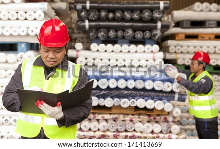 Textile factory worker writing on notepad with partner arranging raw fabrics on background