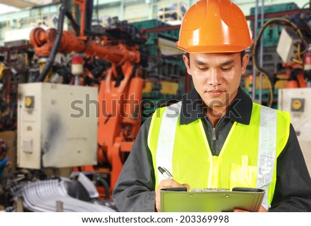 Technician engineer wearing safety vest and yellow helmet writing a notepad, standing in front of robot machine