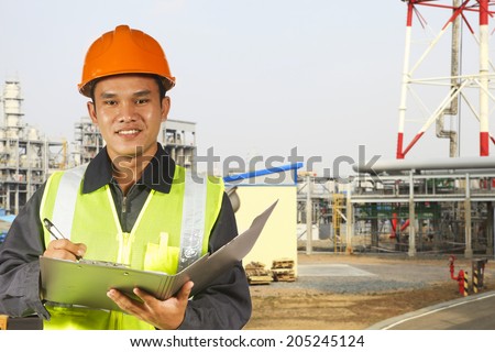 Engineer with a note board and pen in his hands, wearing a hard hat and safety vest in front of a petrochemical plant and refinery