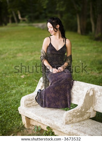 Young woman in long evening gown sitting on the back on a stone park bench