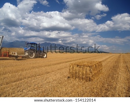 Summer view of harvested wheat field with blue tractor collecting packed crops of hay in Liptov region, central Slovakia. In the distance can be seen peaks of Rohace mountains, the Western Tatras.