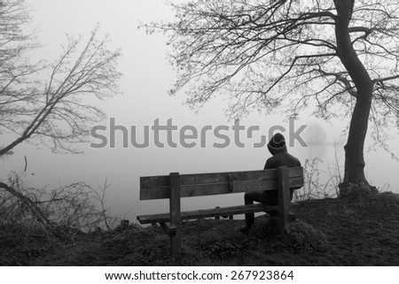 Grieving man sitting alone on a bench at a lake on a foggy day.