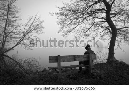 Melancholy emotions concept: sad man sitting on a bench at a foggy lake.